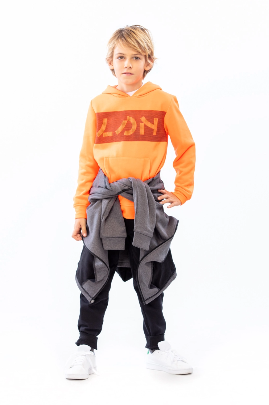 Kid standing looking at camera, with hand on his hip, wearing a orange hoodie with red lettering and a tied jacket on his waist