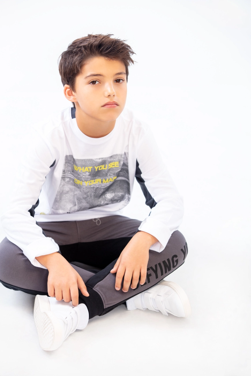 Kid sitting on floor looking at camera, wearing a white shirt with printed image and grey sweat pants
