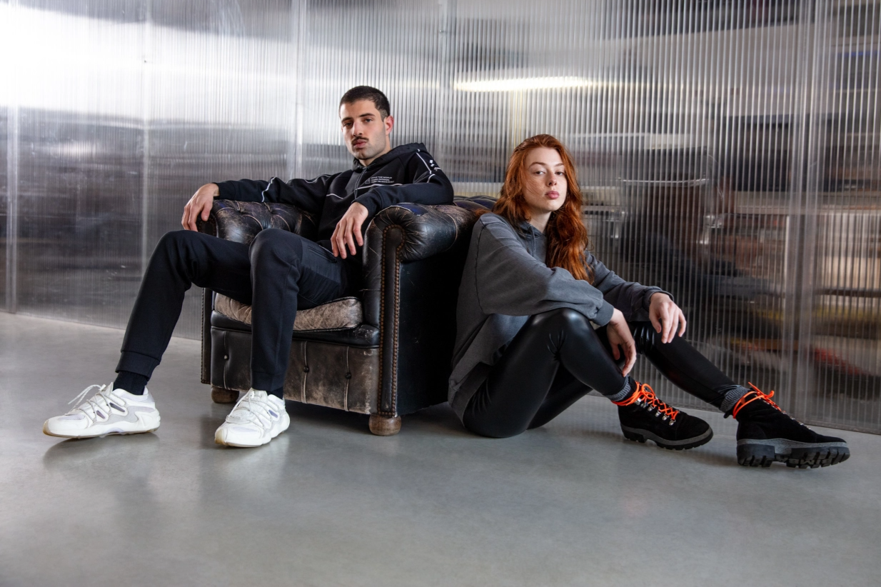 Man sitting on couch wearing a black hoodie next to a woman sitting on floor wearing grey sweatshirt