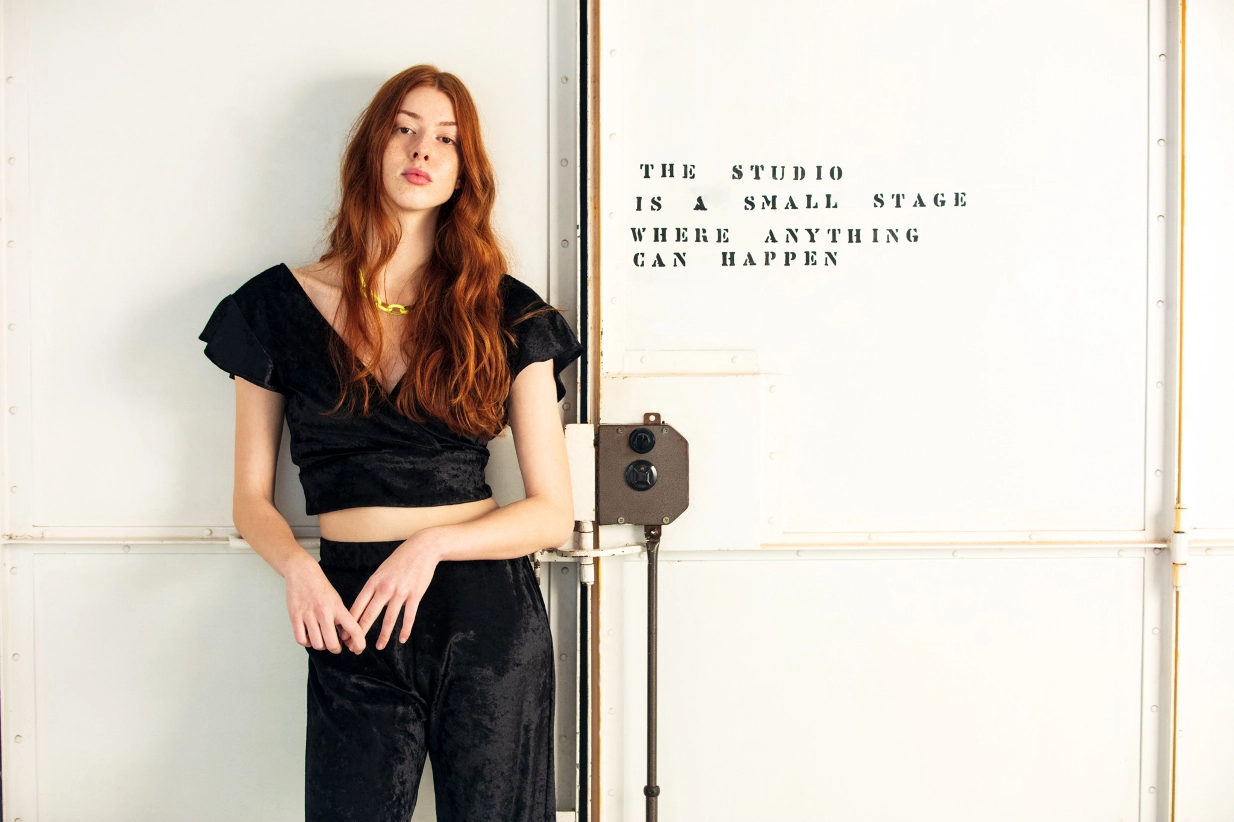 Woman standing up next to a closed door with black setence written into it, wearing a black top and black pants