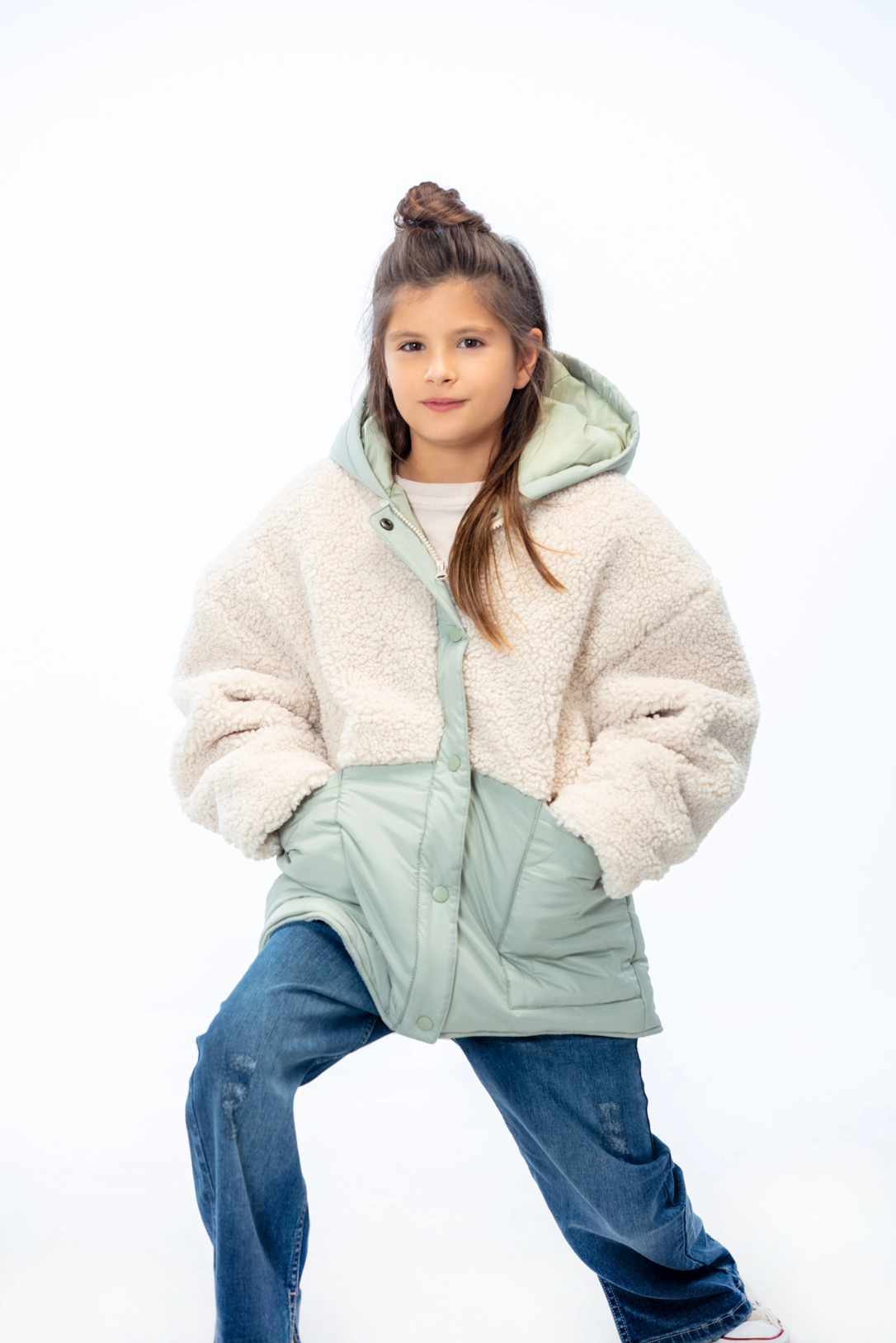 Kid standing, looking at camera wearing a white and green jacket with hands inside pockets and jeans