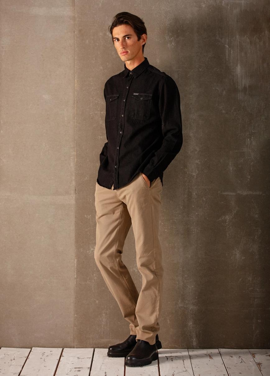 Man leaning against wall looking at camera, wearing a button up black shirt and beige pants