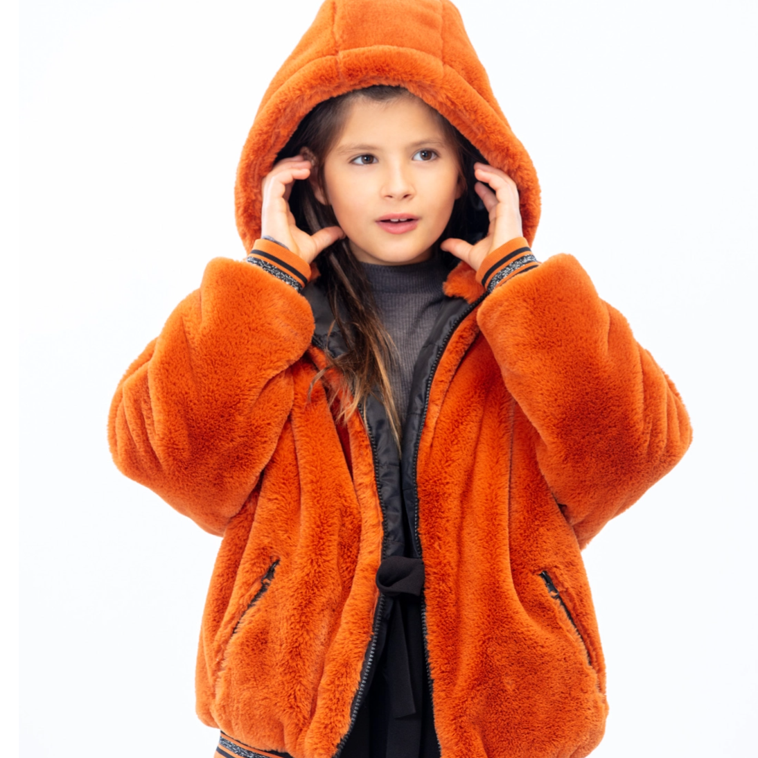 kid standing up looking away from camera, wearing an orange jacket with her hands over her ears