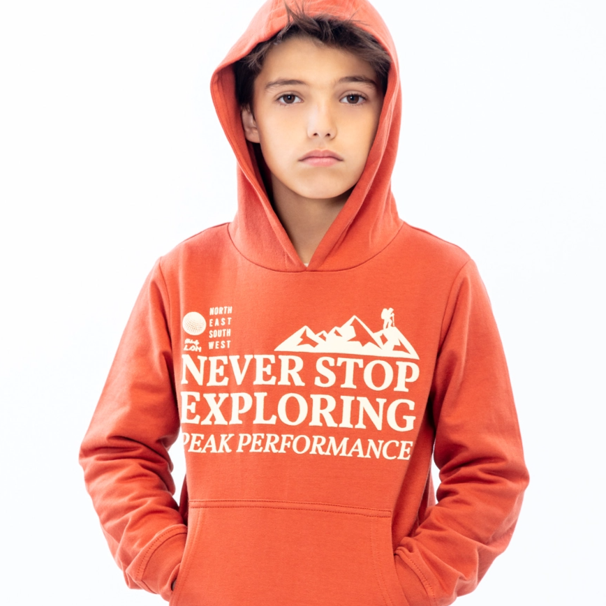 LOSAN- Collection Ground, kid standing up looking at camera, wearing a orange hoodie covering his head and hands in pocket