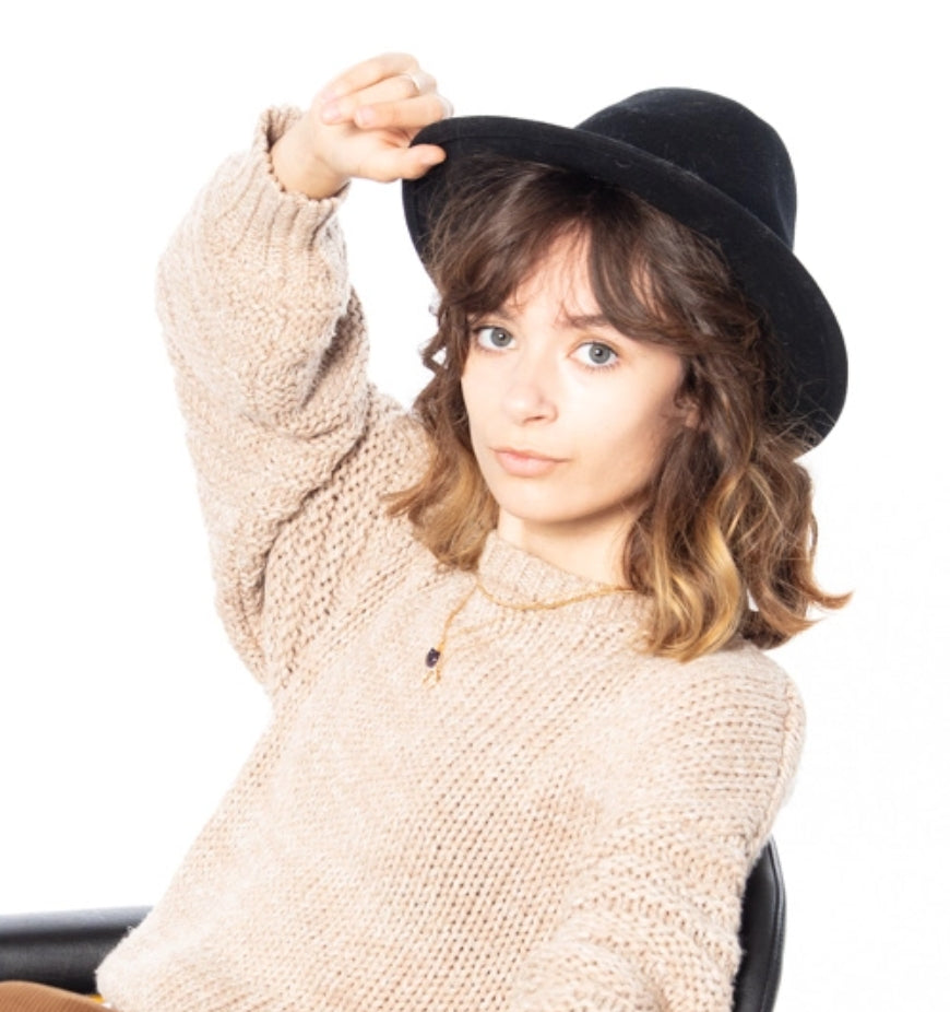 portrait of a woman looking at camera, wearing a beige sweat while holding a black hat