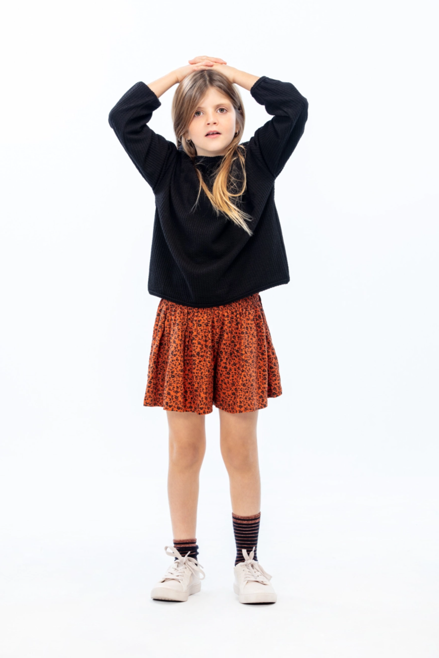 LOSAN- Collection Natural cooper, kid standing up with hands on top of her head, wearing a black sweat and a printed skirt