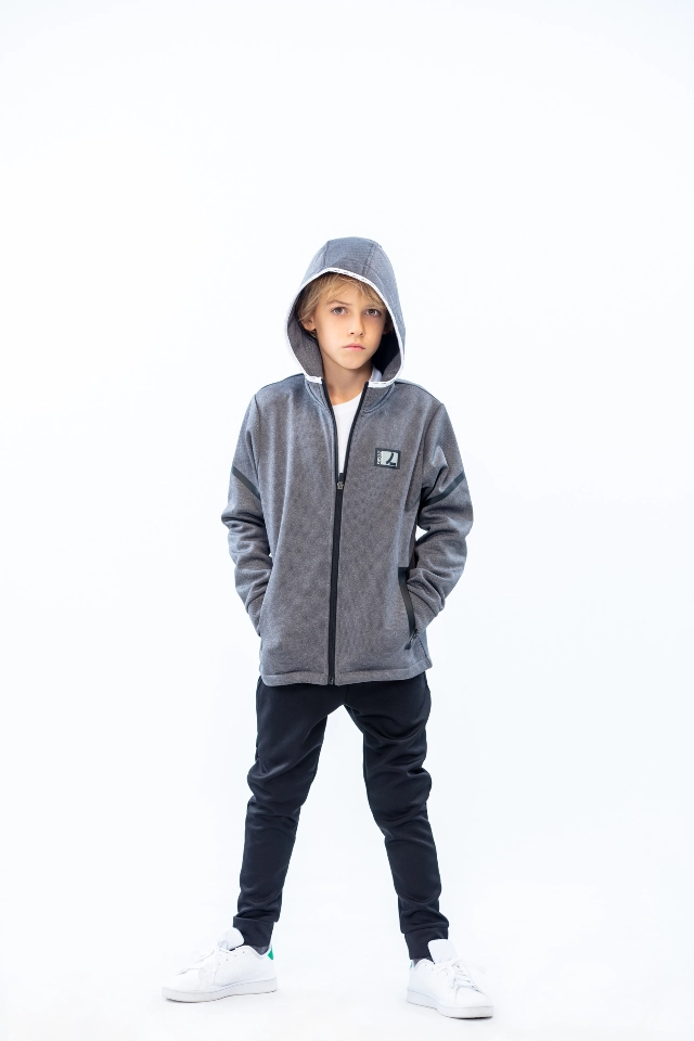 Kid standing up looking at camera wearing a zipped up hoodie with his head covered and hands in pockets