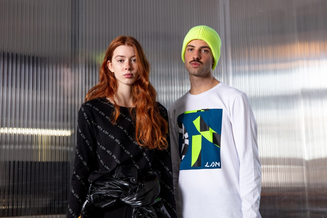 Man wearing a black shirt standing up next to a man wearing a white sweat with blue print wearing a light green beanie