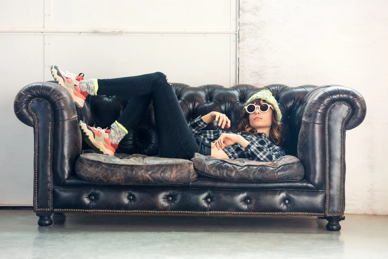 Woman lying on black couch wearing black pants, with plaid shirt, sunglasses and beanie hat