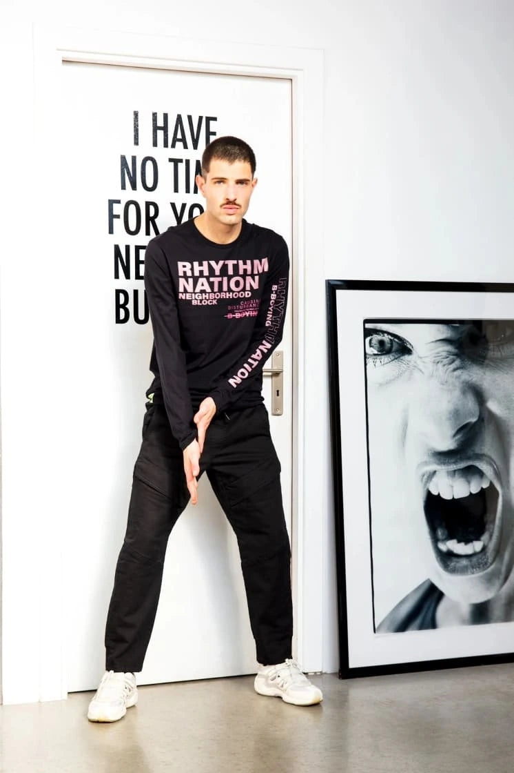 Hombre de pie apoyado en la puerta, vestido con una camiseta estampada, junto a una fotografía enmarcada