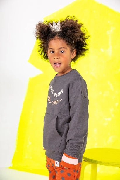 Side profile of a kid standing up looking at camera wearing a crown and a grey sweat with multi colored lettering