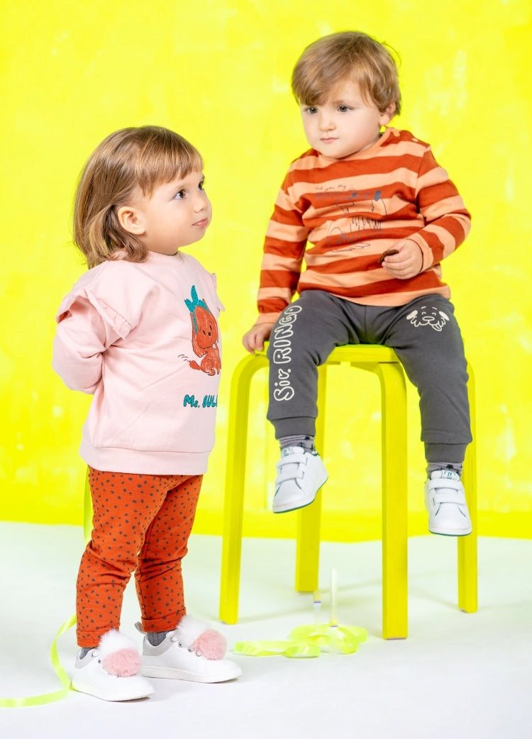 Two toddlers sitting on bench wearing a striped orange and yellow sweat and standing wearing a pink sweat