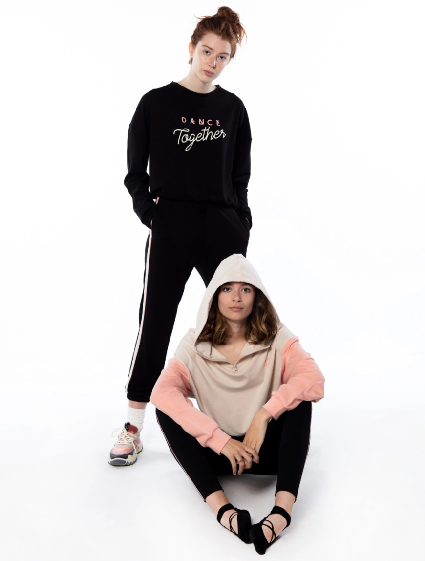 woman standing up wearing a black hoodie with lettering with another woman sitting in front wearing a pink and white hoodie