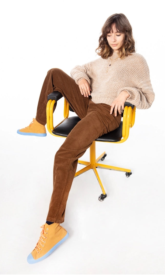 Woman sitting on yellow chair, looking down, wearing white sweat and brown pants and orange sneakers