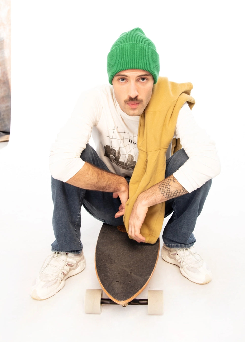 LOSAN - Collection Casual, uomo seduto su skateboard che indossa un berretto verde sudore bianco e una felpa con cappuccio gialla sulla spalla