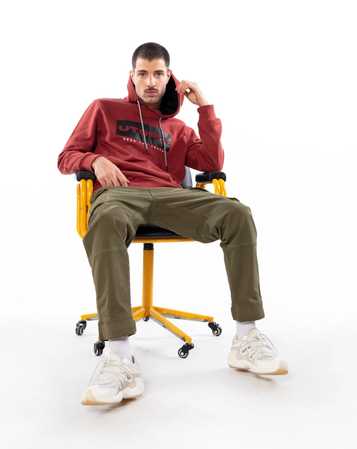 Man sitting on yellow chair, wearing red hoodie with lettering and brown pants